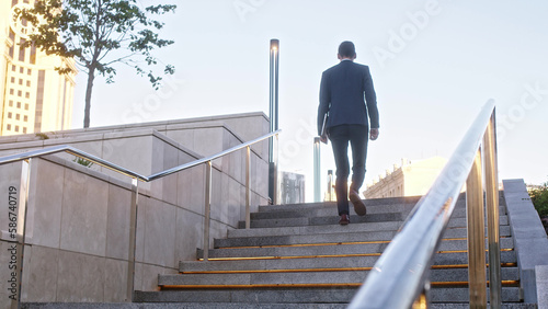 Unrecognizable young businessman climbs up backlit stone stairs in city. man in formal suit dark blue color with tablet in his hands walks forward, close up view of his feet in brown leather boots.