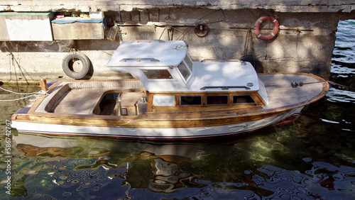 small motor boat is moored to shore of concrete wall. fishing boat is bobbing on waves in bay or boat parking lot. Single-deck water craft with roof.