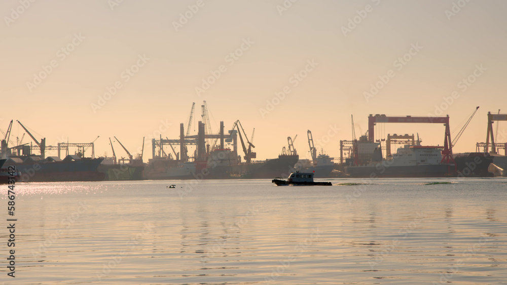 small white coast guard motorboat sails through bay with large port in background with cargo tankers. Sunset or dawn over port city in summer.