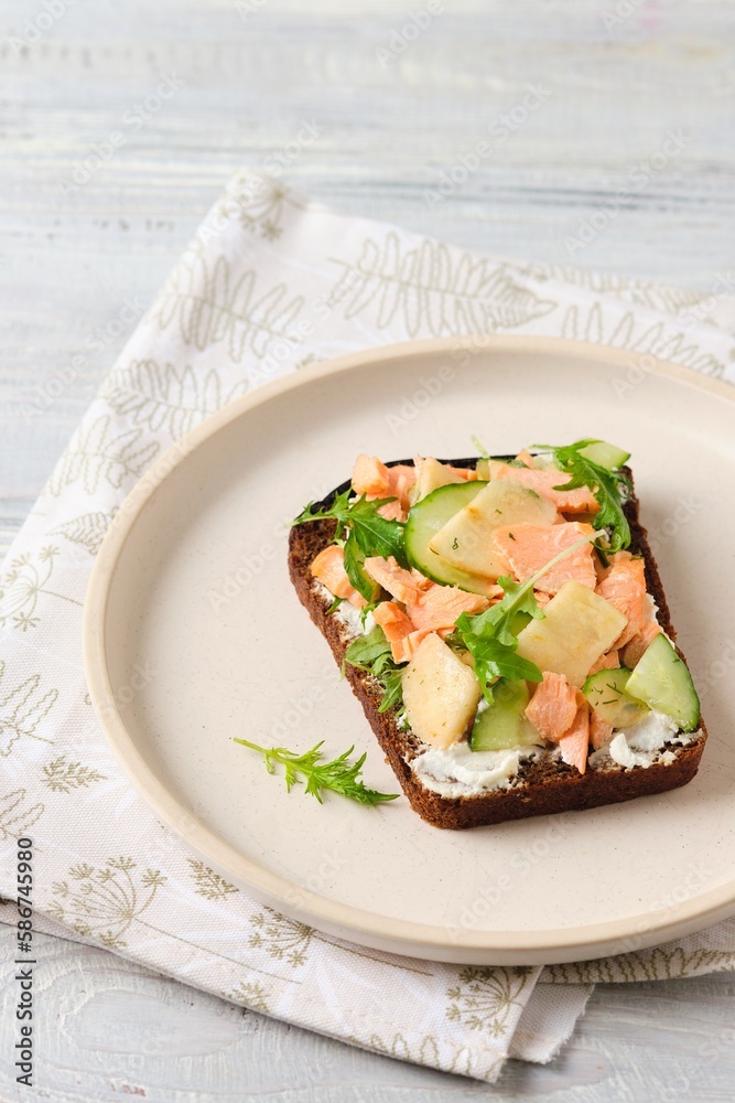 Smorrebrod, an open sandwich on rye bread with boiled salmon, fresh cucumber, cream cheese, apple and herbs on a ceramic plate on a light concrete background. Danish cuisine.