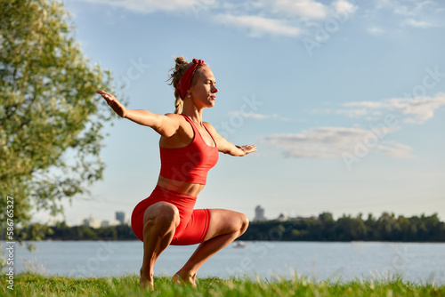 A woman is engaged in animal flow gymnastics, close-up outdoors near the river on green grass in red clothes.