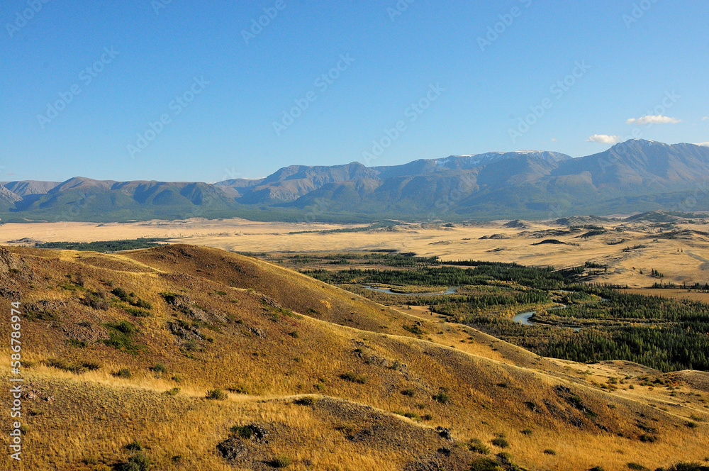 A view from the top of the hill to a valley yellowed by autumn at the foot of a mountain range with snow-capped peaks and a flowing meandering river below.