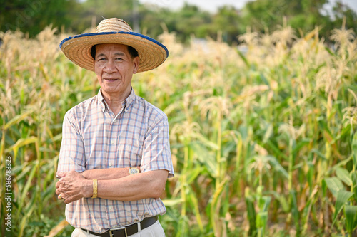 Successful Asian-aged male farmer in straw hat stands in his cornfield with arms crossed.