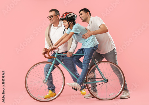 Young man with his father teaching little boy to ride bike on pink background