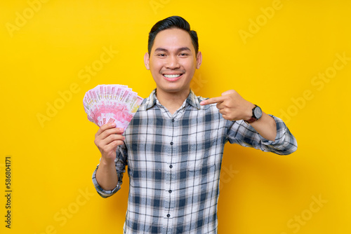 Portrait of smiling young Asian man wearing plaid shirt pointing finger at cash money in rupiah banknotes isolated on yellow background. people lifestyle concept