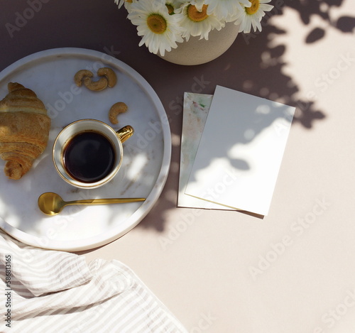 Card mockup in breakfast still life scene with copy space.Cup of coffee, croissant , daisy flowers in a vase top view on beige desk with sunlight shadows. Feminine lifestyle composition. 