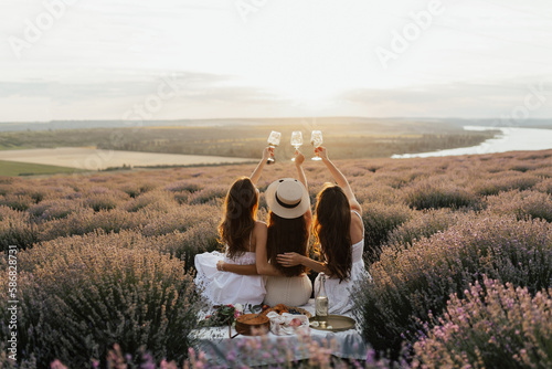 Obraz na płótnie Back view of company of young female friends having fun, drink wine, and enjoy lavender field and sunset at summer picnic. w salonie
