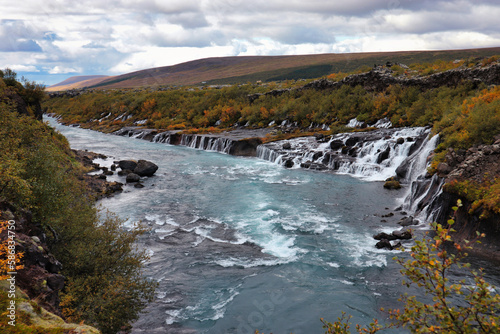 A stunning view of Hraunfossar waterfall in Iceland, where clear waters cascade over lava rocks into a turquoise river, surrounded by autumnal trees and a peaceful landscape photo