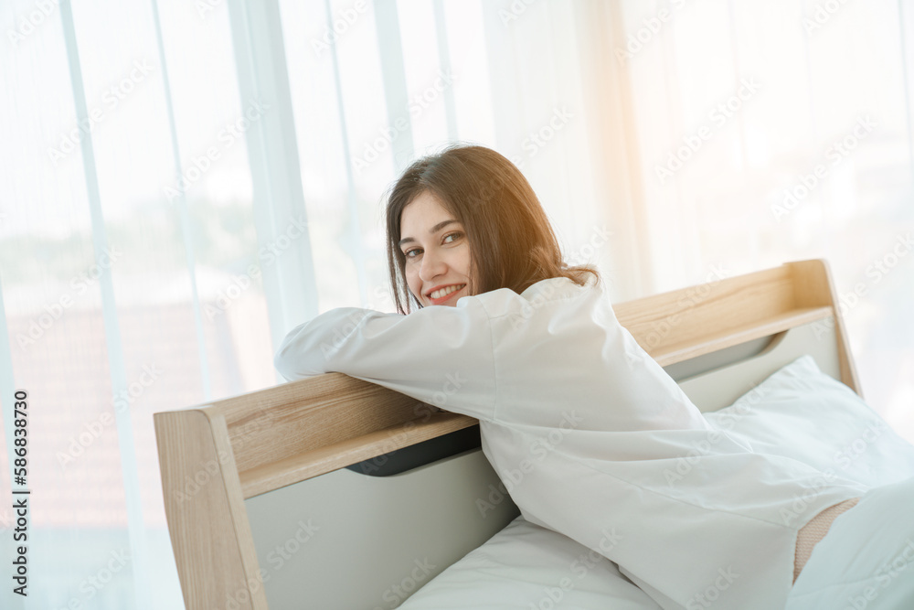 Photo of young happy woman in pajama stretching her arms and smiling while sitting on bed after sleep