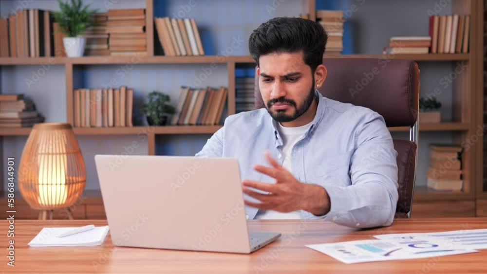 An exhausted and anxious Indian man is having difficulty completing his financial report in office. Man staring at his laptop, surrounded by various charts, graphs on the table. Tension and stress. An exhausted and anxious Indian man is having difficulty completing his financial report in office. Man staring at his laptop, surrounded by various charts, graphs on the table. Tension and stress.