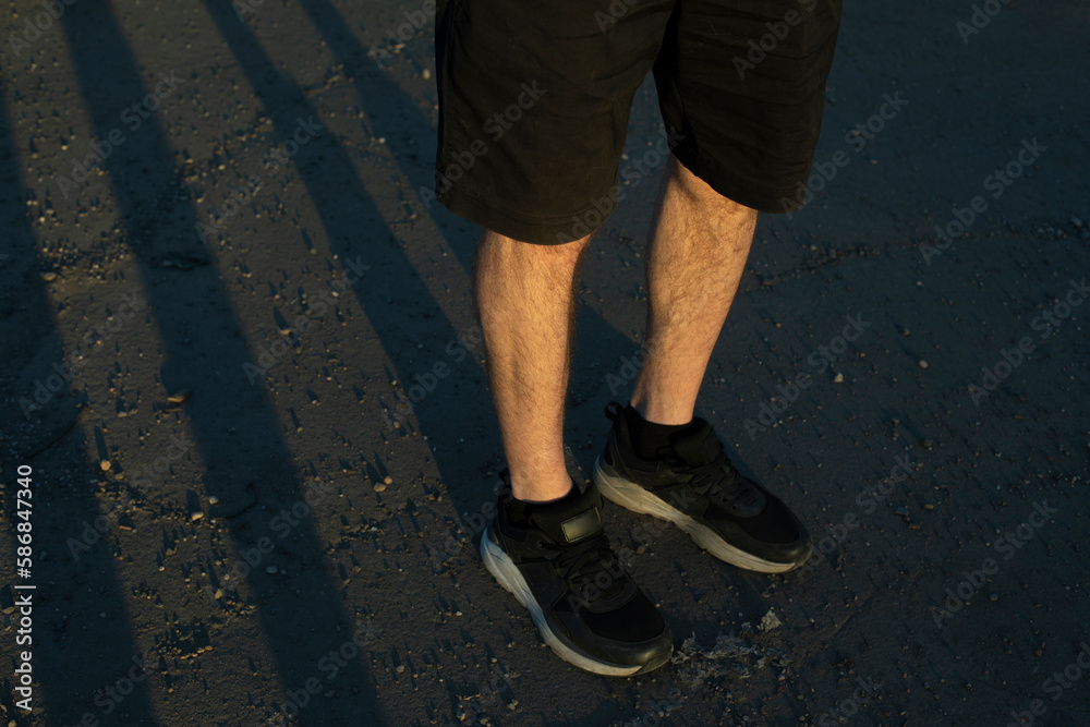 Legs of guy in black shorts and black sneakers. Persons feet stand on ass. Shorts in fashion. Legs of guy in black shorts and black sneakers. Persons feet stand on ass. Shorts in fashion.