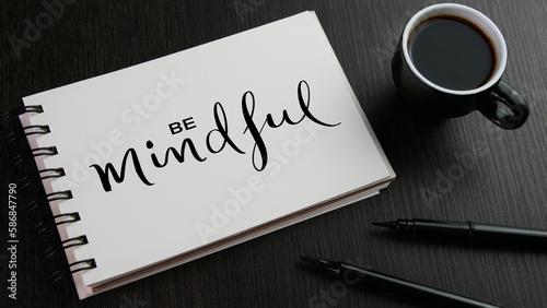 BE MINDFUL black lettering in notebook with cup of coffee and pens on black wooden desk photo