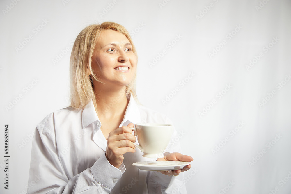 Portrait of pretty blonde smiling woman posing with cup of tea or coffee on white background. Happy girl model in white shirt in studio. The concept of pleasant morning at home or at work. Copy space Portrait of pretty blonde smiling woman posing with cup of tea or coffee on white background. Happy girl model in white shirt in studio. The concept of pleasant morning at home or at work. Copy space