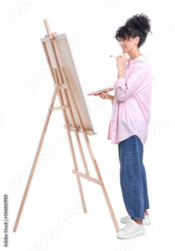 Young woman holding brush and artist`s palette near easel with canvas against white background