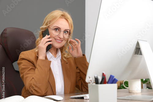 Lady boss talking on smartphone near computer at desk in office. Successful businesswoman