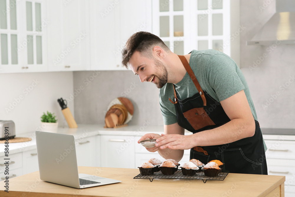 Man decorating muffins with powdered sugar while watching online cooking course via laptop in kitchen. Time for hobby Man decorating muffins with powdered sugar while watching online cooking course via laptop in kitchen. Time for hobby