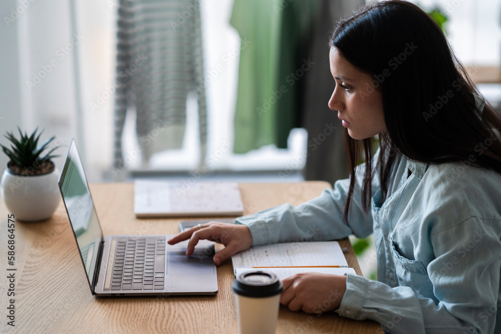 Side view of young female freelancer in casual clothes sitting at table with laptop and cup of coffee while working on project Side view of young female freelancer in casual clothes sitting at table with laptop and cup of coffee while working on project