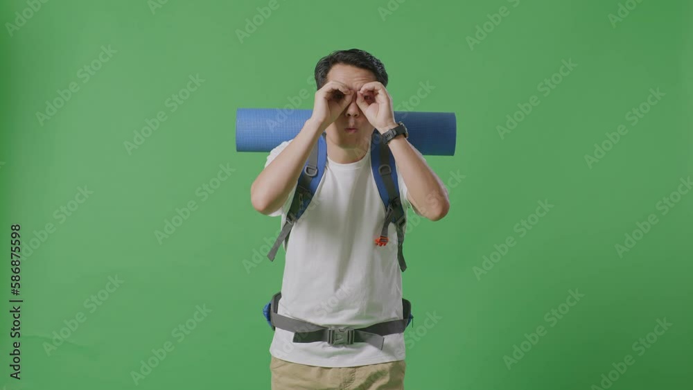 Asian Male Hiker With Mountaineering Backpack Making Binoculars Gesture Looking At Something Then Saying Wow While Standing On Green Screen Background In The Studio
