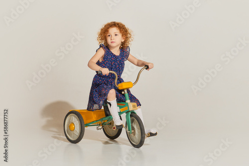 Charming cute girl with curly hair and wearing pattern dress riding retro bike and looking away over light background