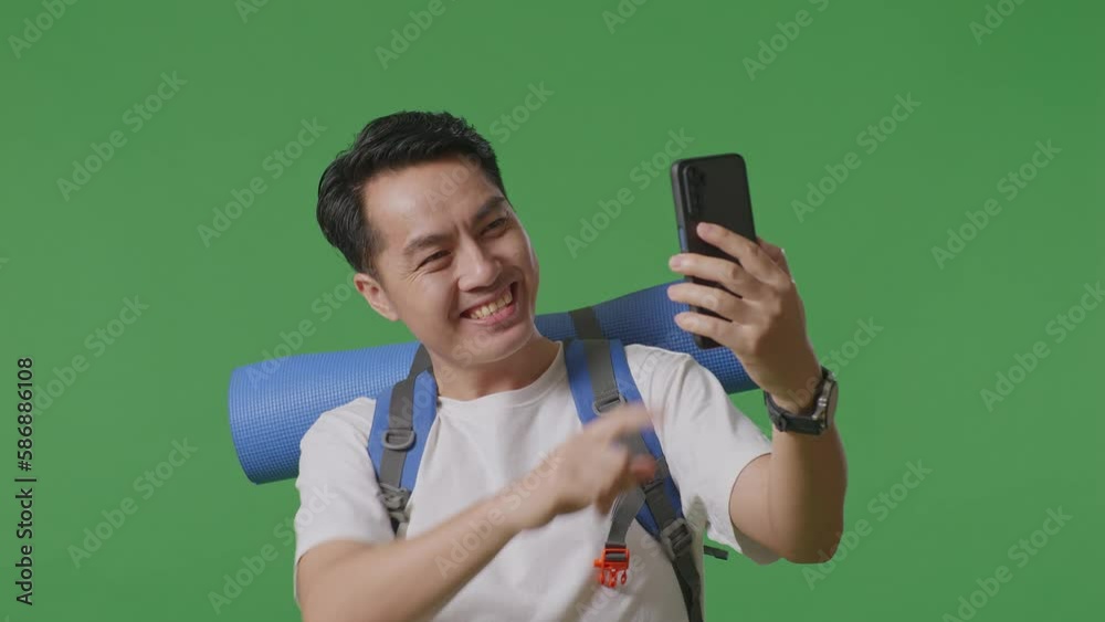 Close Up Of Asian Male Hiker With Mountaineering Backpack Showing Peace And Thumbs Up Gesture While Taking A Selfie On Green Screen Background In The Studio
