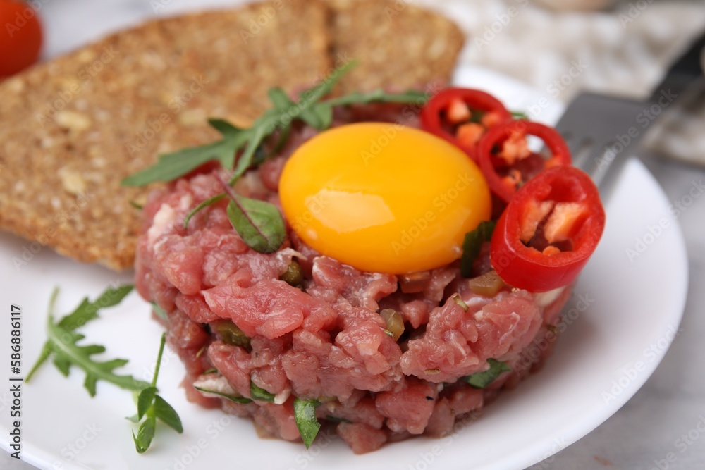Tasty beef steak tartare served with yolk, pepper, bread and greens on white table, closeup Tasty beef steak tartare served with yolk, pepper, bread and greens on white table, closeup