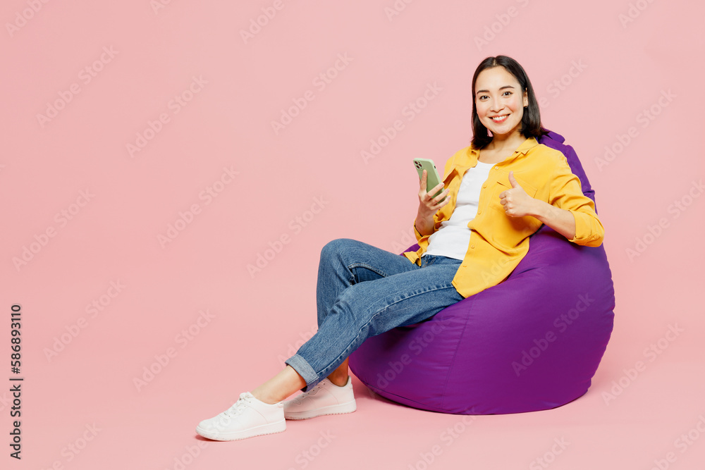 Full body young fun woman of Asian ethnicity wears yellow shirt white t-shirt sit in bag chair hold in hand use mobile cell phone show thumb up isolated on plain pastel light pink background studio. Full body young fun woman of Asian ethnicity wears yellow shirt white t-shirt sit in bag chair hold in hand use mobile cell phone show thumb up isolated on plain pastel light pink background studio.