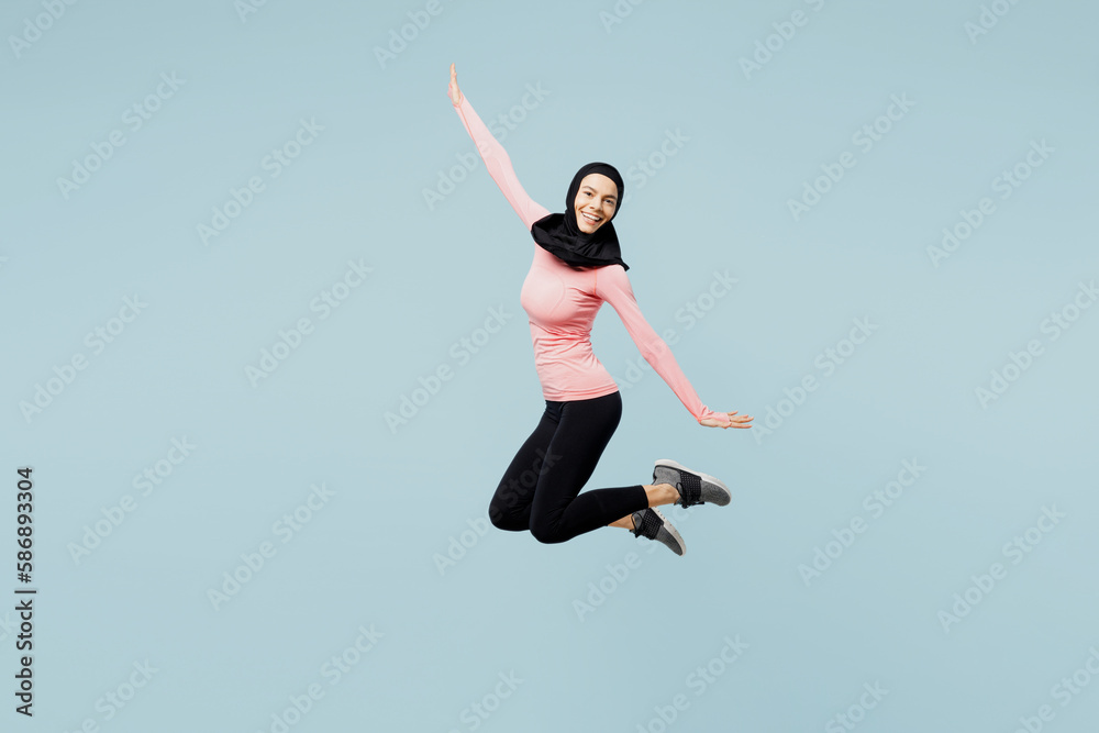 Full body young asian muslim fitness trainer sporty woman wear pink abaya hijab spend time in home gym jump high spread hands isolated on plain blue background studio. Workout sport fit abs concept.