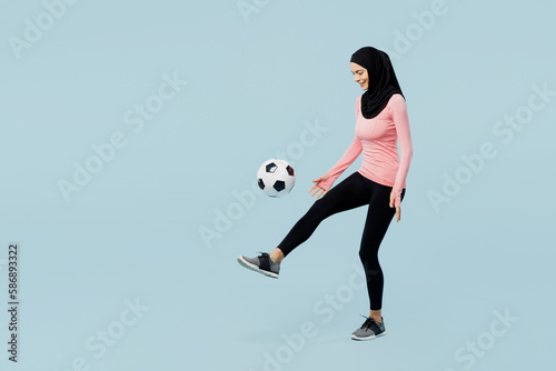 Full body young asian muslim fitness trainer sporty woman wear pink abaya hijab spend time in home gym kicks soccer ball on foot isolated on plain blue background studio Workout sport fit abs concept © ViDi Studio