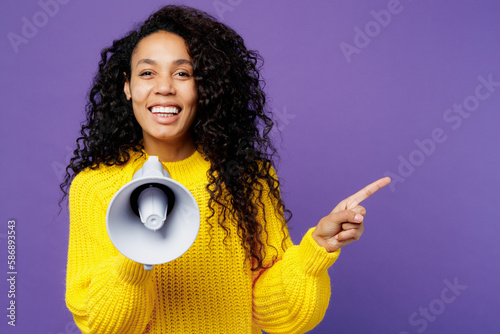 Young fun woman of African American ethnicity wear casual yellow sweater hold in hand megaphone scream announces discounts sale Hurry up isolated on plain purple background studio. Lifestyle concept.