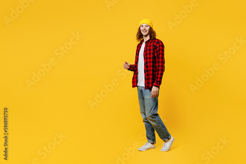Full body sideways smiling happy fun young man wearing red checkered shirt white t-shirt hat walking going strolling look camera isolated on plain yellow background studio portrait. Lifestyle concept.