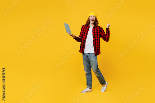 Full body young smiling happy caucasian IT man wearing red checkered shirt white t-shirt hat hold use work on laptop pc computer do winner gesture isolated on plain yellow background studio portrait.