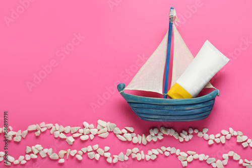 Suntan cream in toy sailboat and white marble pebbles on pink background, flat lay. Space for text
