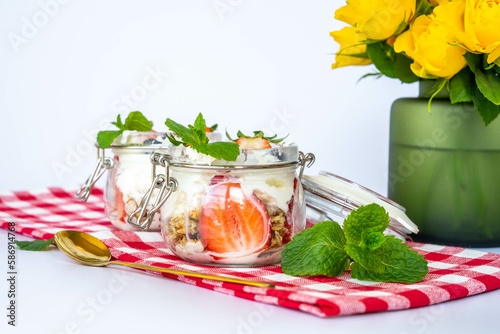 Healthy homemade breakfast with muesli, yogurt and strawberries in small glass jars, on a red and white  squares towel , near yellow roses in a green vase photo