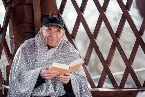 Senior man wrapped in warm wool plaid reads book sitting in wooden summer house outdoor. © Bonsales