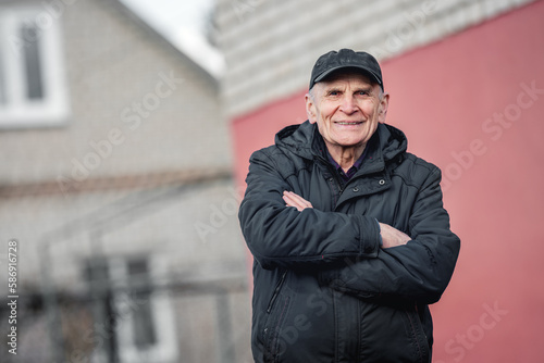 Outdoor personality connected portrait of candid elderly man wearing cap and standing against blurred cottage house background.