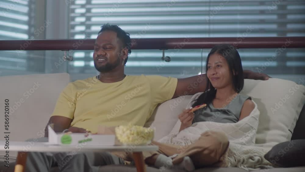 Multiethnic family couple sitting together on sofa at home, having snacks, chatting and smiling while watching TV show together in evening Multiethnic family couple sitting together on sofa at home, having snacks, chatting and smiling while watching TV show together in evening