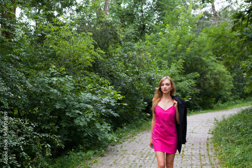 Fototapeta Naklejka Na Ścianę i Meble -  A slender young girl with long blond hair, in a short stylish pink dress, walks beautifully along the path in the forest in summer. She is holding a black jacket on his shoulder.