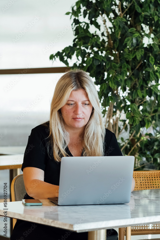 Young blond woman working with laptop while sitting in cafe indoors.