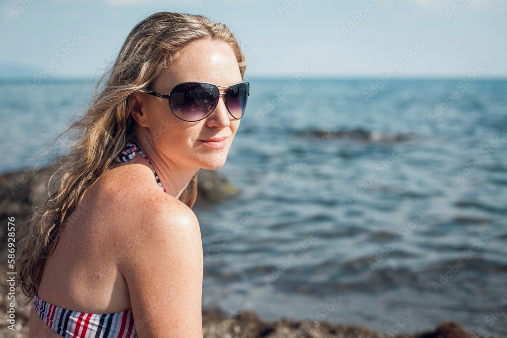 Portrait of a blonde girl in a striped swimsuit and glasses sits on the ocean, on a bright sunny day. Looks into the distance. Portrait of a blonde girl in a striped swimsuit and glasses sits on the ocean, on a bright sunny day. Looks into the distance.