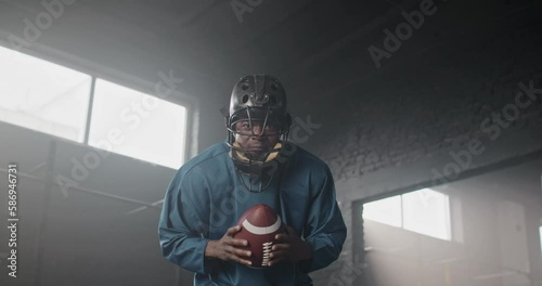 Attractive African American man throwing ball up and looking straight into camera in dark room. Handsome male with helmet and wearing sportswear getting ready for game. American football concept photo