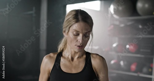 Portrait of Caucasian woman wearing fitness top and having boxing gloves, standing in front of camera in dark background of sports studio. Pretty female looking exhausted after training heavily. photo