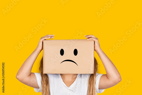 Mujer con caja de cartón en la cabeza con gesto pensativo preocupante y manos sosteniendo la cabeza sobre fondo amarillo liso brillante y aislado. Vista de frente y de cerca. Copy space