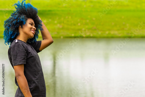 Uma jovem negra, feliz, num parque natural de muito verde, alisando os cabelos tingidos de azul.