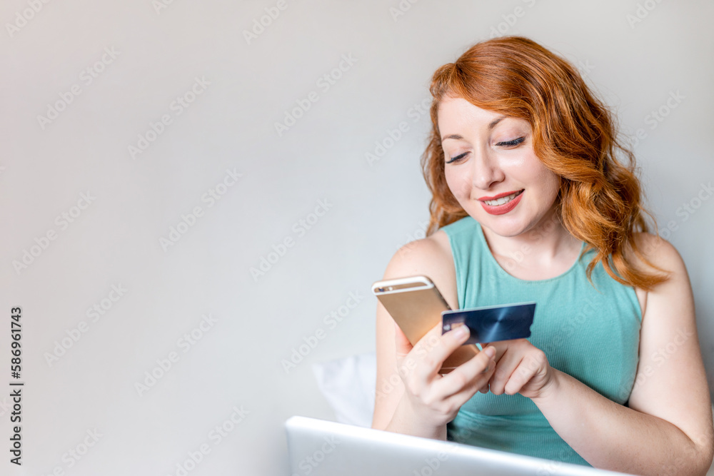Young Caucasian redhead girl using smartphone and credit card in front of laptop in bed at home with copy space and grey background Young Caucasian redhead girl using smartphone and credit card in front of laptop in bed at home with copy space and grey background