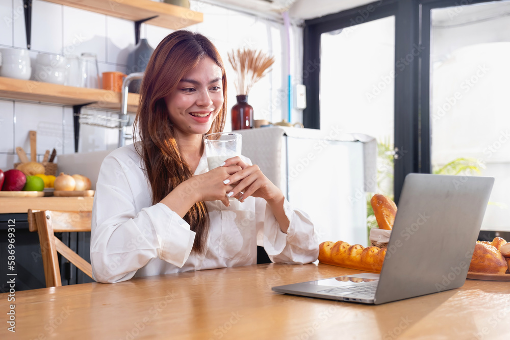 Woman with a beautiful face in a white shirt is making a healthy breakfast with bread, vegetables, fruit and milk inside the kitchen and opening her laptop for cooking lessons. healthy cooking ideas. Woman with a beautiful face in a white shirt is making a healthy breakfast with bread, vegetables, fruit and milk inside the kitchen and opening her laptop for cooking lessons. healthy cooking ideas.
