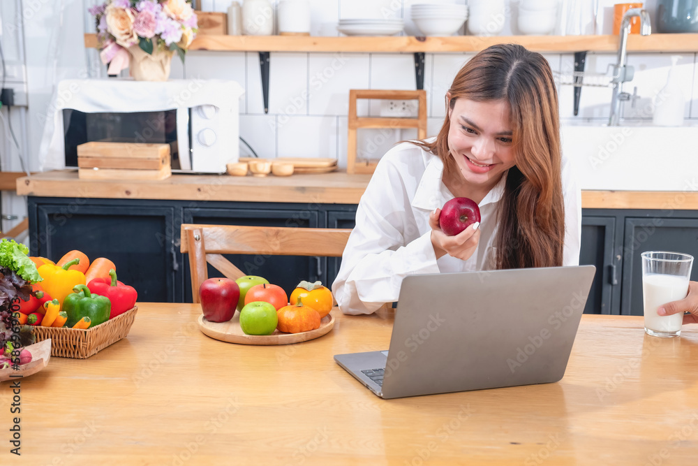 Woman with a beautiful face in a white shirt is making a healthy breakfast with bread, vegetables, fruit and milk inside the kitchen and opening her laptop for cooking lessons. healthy cooking ideas.