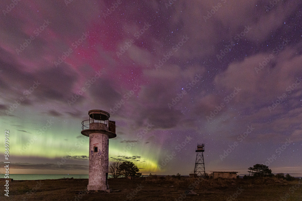 Coastal night scenery with the abandoned old lighthouse and Soviet military watchtower in the intensive aurora colored sky and sea background