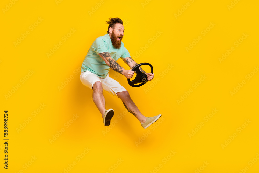 Full body cadre of overjoyed young jump man red hair wear green t-shirt hold steering wheel driving fast speed new car isolated on yellow background