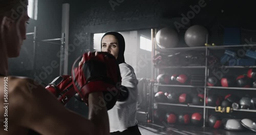 Beautiful females practicing punches in gym together. Attractive Caucasian woman actively training power exercises with boxing gloves while sparring coach holding sports paws. Sport boxing concept. photo