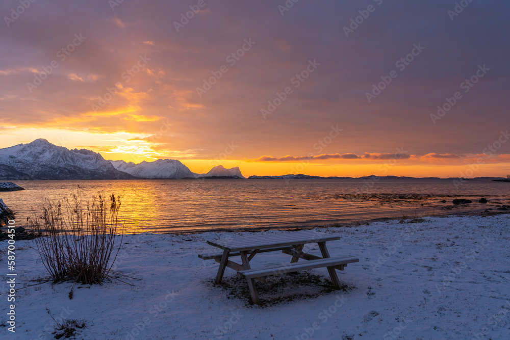 Abendrot mit Brücke, Baum, Rastplatz und den verschneiten Bergen von Senja, Norwegen. Panorama. die roten Wolken spiegeln sich in Wasser des Fjords bei Skaland. Sonnenuntergang am Bergsfjorden.